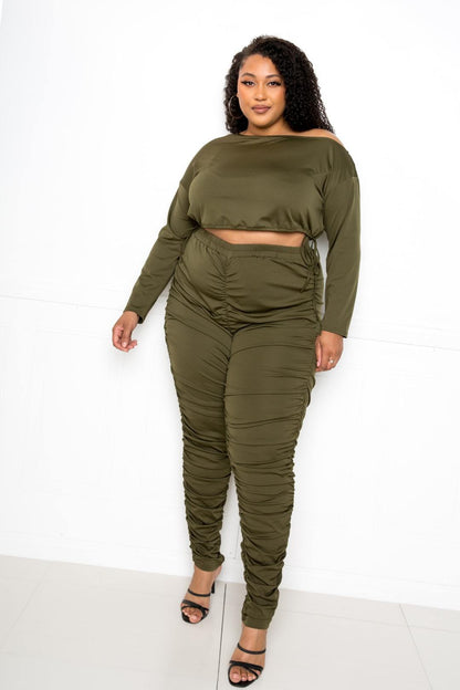 Plus-size model wearing an olive green off-shoulder cropped top and ruched leggings set, designed for curvy figures, standing indoors.